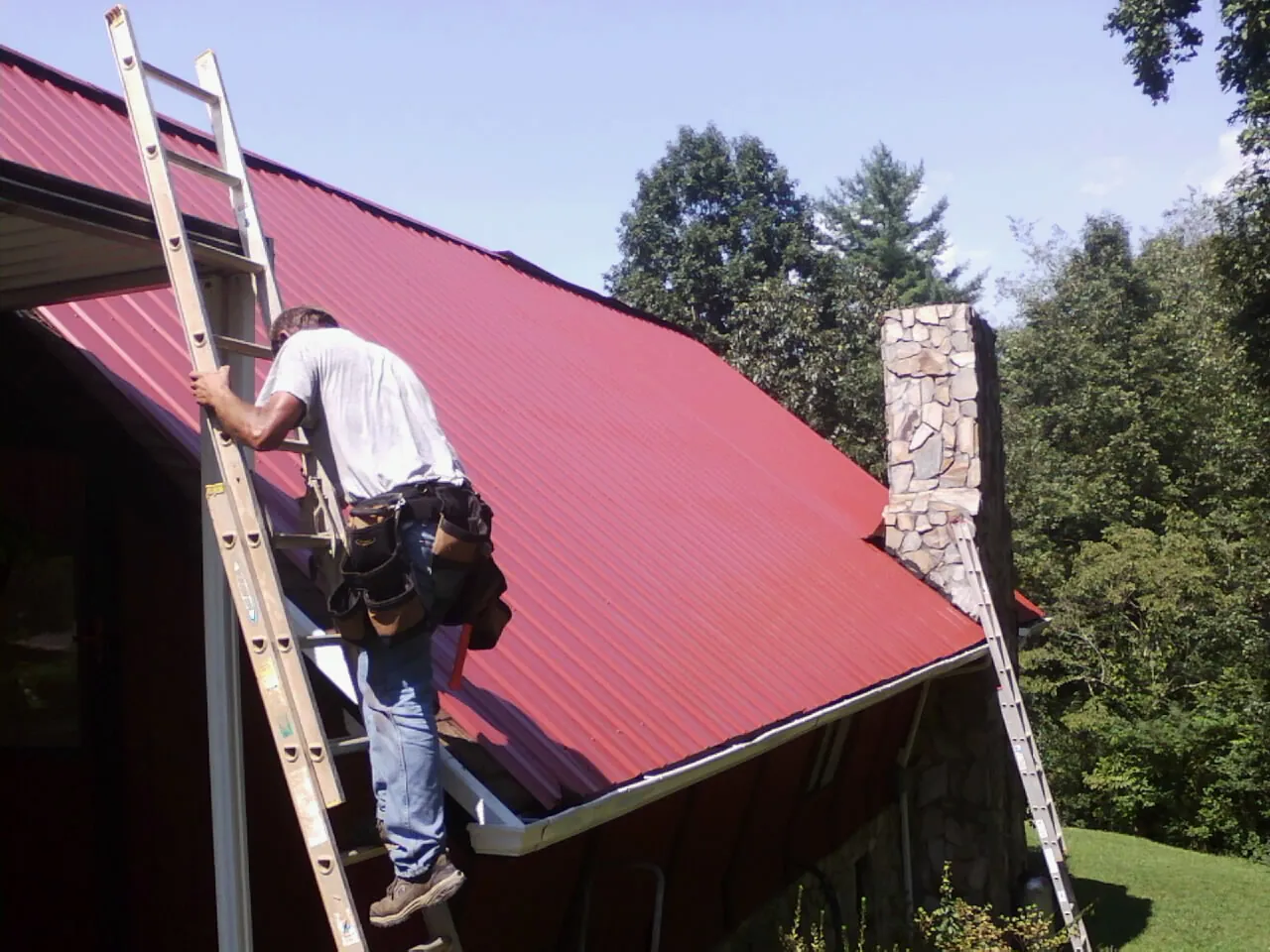 Roofer on a ladder beside a red metal roof and stone chimney — Soffit & Fascia Repair by Roofing Brothers in Lincoln City